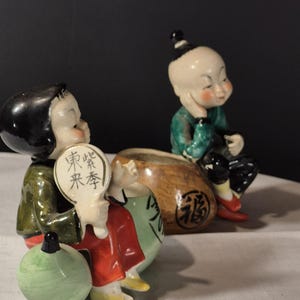 Peut inclure: Deux figurines en céramique représentant des enfants asiatiques. L'une tient un éventail avec du texte, vêtue d'une tenue rouge et verte. L'autre est assise, portant une veste verte et un pantalon noir. Un globe vert et un sac marron sont également présents.