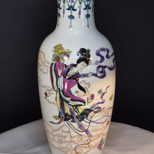 May include: A white porcelain vase with a gold rim and base, featuring a hand-painted design of a woman in flowing robes. The woman is surrounded by clouds and ribbons in shades of purple, pink, and yellow. The vase has a detailed floral pattern at the top.
