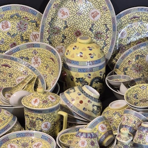 Pode incluir: Uma coleção de pratos de porcelana amarelos e brancos com desenhos florais e caracteres chineses. O conjunto inclui pratos, tigelas, xícaras e um pote com tampa. As bordas têm um padrão geométrico azul e branco.