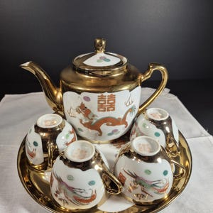 Op de afbeelding: Een met goud afgewerkte theeset met een theepot, kopjes en een dienblad. De witte porseleinen stukken hebben draken- en bloemmotieven in rood, groen en goud. De theepot heeft een gouden handvat en deksel.