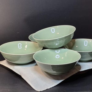May include: Five pale green ceramic bowls with a glossy finish. The bowls are arranged on a white cloth, set against a dark background. The bowls have a simple, elegant design. They are likely for serving food, perhaps 15-20 cm in diameter.