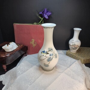 May include: A white ceramic vase with a bird and branch design. The vase has a flared top and a detailed base. A smaller vase with a landscape design is also visible. The background includes a red book and a purple flower.