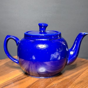 May include: A cobalt blue ceramic teapot with a matching lid. The teapot has a rounded body, a curved spout, and a loop handle. The glossy finish reflects light, highlighting the rich color. The teapot is a classic design for brewing tea.