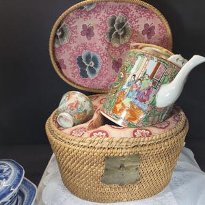 Könnte beinhalten: Ein Vintage-Teeservice mit Teekanne, Tasse und einem Weidenkorb. Teekanne und Tasse sind mit farbenfrohen Szenen verziert. Der Korb hat einen rosa Deckel mit Blumenmuster. Ein charmantes Beispiel für antike Handwerkskunst.
