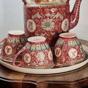 May include: A red and gold ceramic tea set with a teapot and three teacups on a tray. The teapot has a woven handle. The set features floral and geometric patterns and Chinese characters.