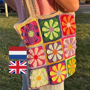 Op de afbeelding: Een handgemaakte gehaakte tote bag met een kleurrijk bloemmotief. De tas heeft een patchwork van vierkanten, elk met een bloem in een andere kleur. De tas is beige met een roze voering en wordt door een persoon vastgehouden.