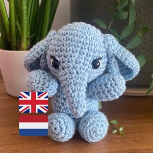 Op de afbeelding: Een lichtblauwe gehaakte olifant met zwarte ogen en grote oren. De handgemaakte amigurumi olifant zit op een houten oppervlak. Een schattig, handgemaakt speelgoed.