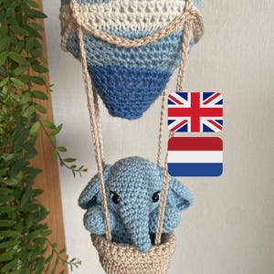 May include: A handmade crochet hot air balloon in blue and cream, with a light brown basket holding a blue elephant. The balloon is suspended by light brown cords. The elephant has black eyes and large ears.