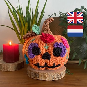 Op de afbeelding: Een oranje gehaakte pompoen met een lachend gezicht en kleurrijke bloemaccenten. De pompoen staat op een houten schijf, met een brandende rode kaars en groen in de achtergrond. Een handgemaakte decoratie.