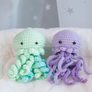 May include: Two handmade crochet jellyfish toys. One is light green with blue tentacles, the other is purple with purple tentacles. Both have black safety eyes and are sitting on a white, fluffy surface.