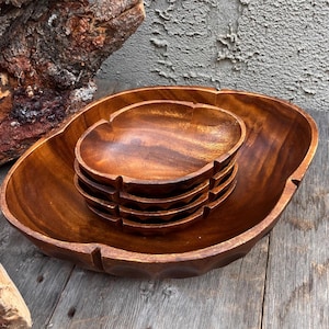 May include: A set of wooden serving bowls in varying sizes. The largest bowl is oval-shaped, with a carved rim. Inside, a stack of smaller, scalloped-edge bowls rests. The wood has a warm, reddish-brown tone, with visible grain.
