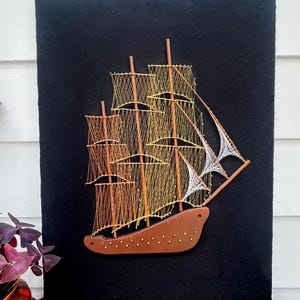 Peut inclure: Une œuvre d'art en fil représentant un voilier. Le voilier est fait de fil marron, jaune et blanc sur un fond noir. La coque et les mâts sont bruns, les voiles sont faites de fil. L'œuvre est montée sur un matériau noir semblable à du feutre.