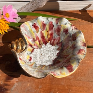 May include: A ceramic seashell-shaped bowl with a gold-coloured spiral handle. The bowl's interior features a pattern of red, yellow, and white brushstrokes. The rim is trimmed with gold. The bowl is approximately 20 cm wide.