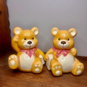 Puede incluir: Dos figuras de osos de peluche de cerámica. Los osos son de color marrón claro con vientres blancos, lazos rosas y pequeños libros. Un oso levanta la pata y el otro sostiene un libro. Los osos tienen ojos negros y narices marrones.
