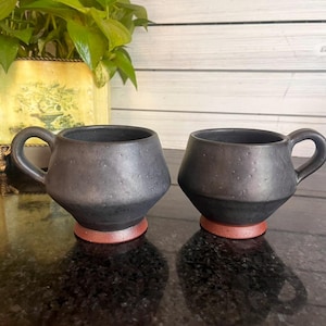 May include: Two black ceramic mugs with a textured surface and a small handle. The mugs have a geometric shape and sit on a reddish-brown base. The mugs are displayed on a dark, reflective surface.