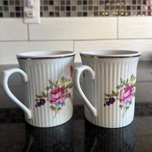 Puede incluir: Dos tazas de cerámica blanca con ribete dorado y diseños florales. Las tazas tienen una textura acanalada y presentan rosas rosas, flores moradas y hojas verdes.