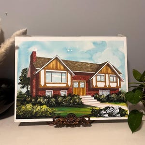 Puede incluir: Pintura a la acuarela de una casa de dos pisos con exterior de ladrillo rojo, tejado marrón y detalles en madera. La casa tiene varias ventanas, una chimenea y está rodeada de arbustos verdes y un cielo azul con nubes. La obra de arte se exhibe en un soporte de metal decorativo.