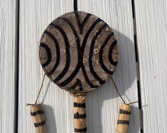 May include: A handmade wooden rattle with a circular head featuring a brown and black patterned design. Two small wooden cylinders with black stripes hang from the sides. The handle is also made of wood with black stripes.