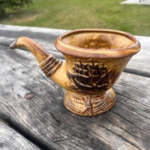 May include: A ceramic tobacco pipe-shaped ashtray in shades of brown and tan. The bowl features a detailed relief of a sailing ship. The pipe has a curved stem and a textured base, evoking a vintage aesthetic.