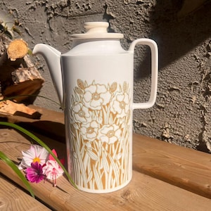 May include: White ceramic coffee pot with a lid and handle. The pot has a gold floral design. It is on a wooden surface. The pot is approximately 25 cm tall.