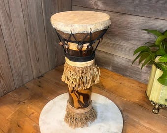 May include: A handcrafted djembe drum with a natural hide drumhead. The drum has a brown wooden body with decorative carvings, wrapped with black and tan rope, and a fringed skirt. It stands on a white marble base, showcasing a unique musical instrument.