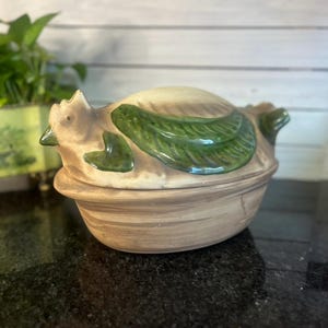 Puede incluir: Un plato cubierto de cerámica con forma de gallina. El plato es principalmente de color marrón con detalles de hojas verdes en la tapa. La gallina tiene una cabeza y una cola detalladas. El plato está sobre una superficie oscura.