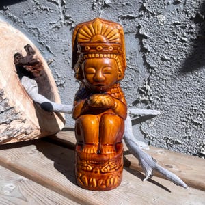 May include: A brown ceramic statue of a seated figure with a detailed headdress and ornate base. The statue has a glossy finish and is posed with hands clasped. The base of the statue has the word "KANZIA" carved into it.