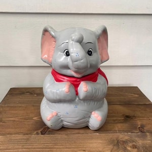 May include: A grey ceramic elephant figurine with pink inner ears and a red scarf. The elephant has blue accents and is sitting on a wooden surface. The figurine is a decorative item.