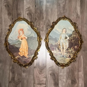 Puede incluir: Dos placas de pared vintage con marcos dorados ornamentados. Cada placa presenta una escena pintada de figuras en un paisaje. Una representa a una mujer con un vestido melocotón, la otra a una persona con atuendo blanco. El fondo muestra montañas y un cielo.