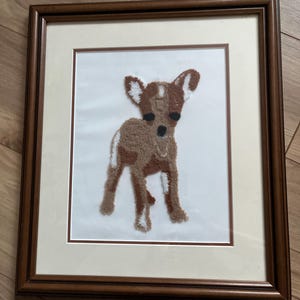 Puede incluir: Una obra de arte enmarcada que presenta un chihuahua marrón y blanco hecho con hilo. El chihuahua está de pie sobre un fondo blanco y está rodeado por un marco marrón.