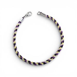 May include: A woven bracelet featuring black, yellow, and purple threads. The bracelet has a silver-coloured clasp and a matching end piece. The bracelet is displayed against a white backdrop.