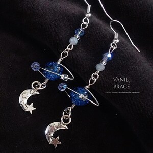 May include: Dangle earrings featuring a silver-tone hook, blue and clear beads, a dark blue glittery planet charm with a silver ring, and a silver crescent moon charm with stars. The text VANIL BRACE Crafted for every era, beaded for every soul is visible.