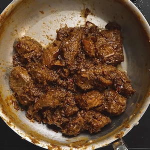 May include: A silver-toned metal skillet containing chunks of cooked, dark brown meat coated in a thick, textured sauce. The meat pieces are irregular in shape and size, showing a deep, savory glaze with small flecks of red and green herbs throughout.