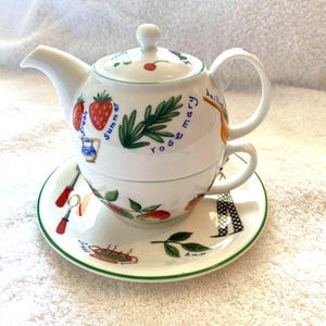 Könnte beinhalten: Weißes Keramik-Teekannen- und Teetassen-Set mit grünem und floralem Design. Die Teekanne hat einen Deckel und einen Griff. Die Teetasse hat einen Griff. Das Set zeigt eine Vielzahl von Kräutern und Früchten, darunter Rosmarin, Erdbeeren und Lorbeerblätter. Der Text "fresh summer rosemary boiling" ist auf die Teekanne gedruckt.