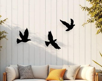 May include: Three black metal bird silhouettes are mounted on a white wooden fence. The birds are in flight, creating a sense of movement. The scene is set in an outdoor living space with a sofa, rug, and plants.