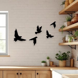 May include: Black metal bird silhouettes in various flying poses are mounted on a white brick wall. The kitchen setting includes wooden cabinets, a white sink, and open shelves with plants and decor. A window is visible.