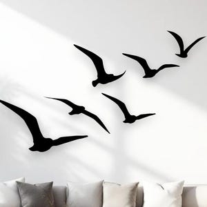 May include: A set of six black bird silhouettes arranged in a flying formation on a plain white wall. The birds are depicted in various poses with wings spread, creating a dynamic visual effect across the wall surface above a sofa.
