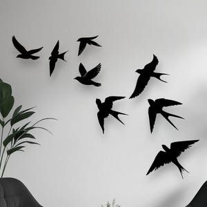 May include: A set of eight black bird wall decals arranged in a flying formation on a plain white wall. The silhouettes depict birds with spread wings and forked tails, creating a dynamic visual effect of a flock in motion across the wall surface.