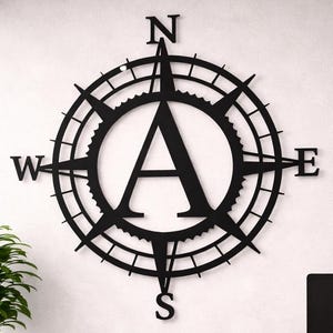 May include: Black metal compass rose wall art with the letter "A" in the center. The compass points North, South, East, and West. The wall art is mounted on a white wall above a wooden desk.