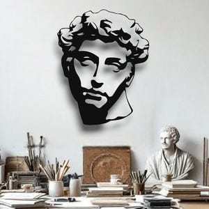 May include: A black metal wall art piece depicting a classical male bust, mounted on a white wall. The artwork is the focal point, with other classical busts and a work table in the background. The room is well-lit with a hanging lamp.