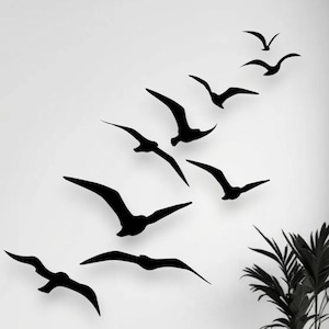 May include: A set of black wall decals featuring a flock of nine birds in flight, arranged in an upward diagonal pattern. The silhouettes depict birds with wings spread in various positions, creating a sense of movement across the white wall.