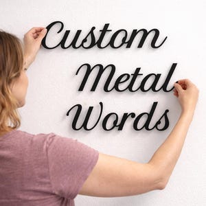 Könnte beinhalten: Schwarze Metallwörter mit der Aufschrift "Custom Metal Words" werden an einer weißen Wand angebracht. Die Wörter sind in einer Schreibschrift. Die Person, die die Wörter platziert, trägt ein rosa T-Shirt.