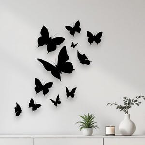 May include: A collection of twelve black butterfly wall decals arranged in a vertical cluster. The butterflies vary in size and wing shape, creating a dynamic visual effect against the plain white wall above a white sideboard.