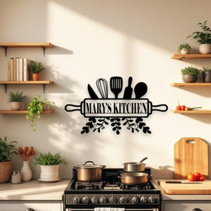 Può includere: Insegna da cucina in metallo nero con la scritta "MARY'S KITCHEN" in un design a matterello, con utensili da cucina e decorazioni a foglie. L'insegna è montata su una parete bianca sopra un fornello.