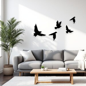 May include: Black metal bird wall art in flight, displayed on a white wall above a gray sofa. The birds are various sizes and shapes, creating a dynamic and modern home decor accent. A palm plant in a woven basket and a wooden coffee table complete the scene.