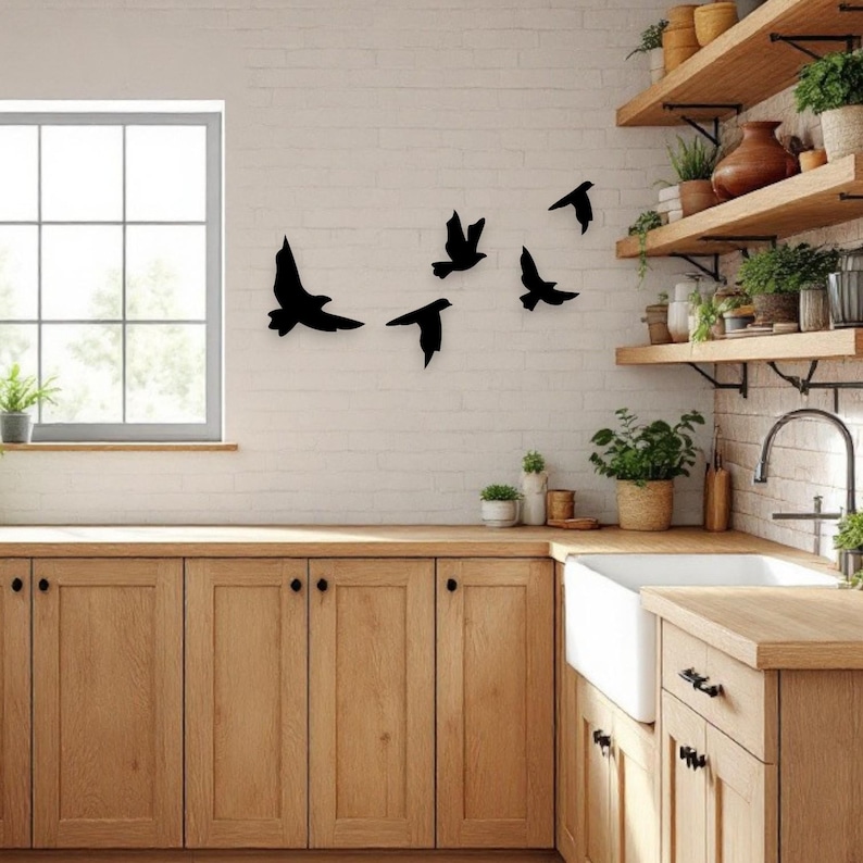 May include: Black metal bird silhouettes in various flying poses are mounted on a white brick wall. The kitchen setting includes wooden cabinets, a white sink, and open shelves with plants and decor. A window is visible.