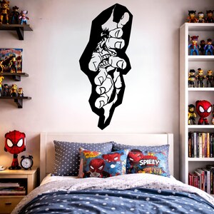 May include: A black and white wall decal depicting a comic book character's hand reaching out. The decal is above a bed with blue and white bedding and Spider-Man themed pillows. Shelves with toys and books are visible.
