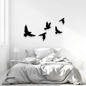 May include: A bedroom scene featuring a bed with a gray comforter and white pillows. Above the bed, five black bird silhouettes are mounted on the white wall. A small white side table with a lamp is on the left.