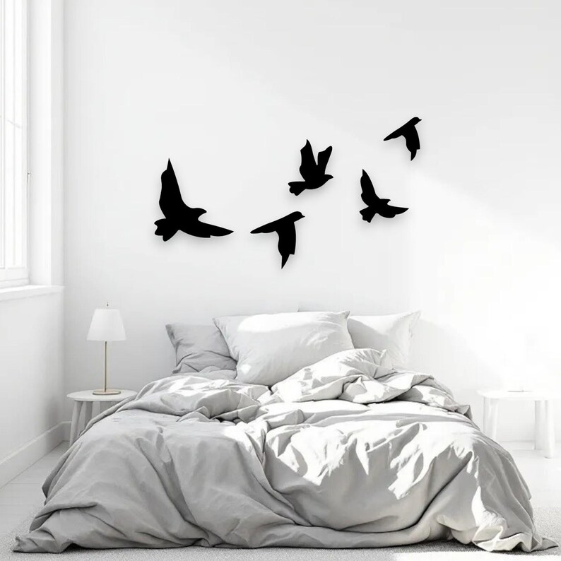 May include: A bedroom scene featuring a bed with a gray comforter and white pillows. Above the bed, five black bird silhouettes are mounted on the white wall. A small white side table with a lamp is on the left.