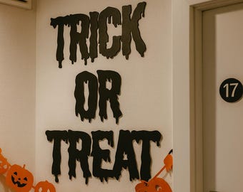 May include: Black wall lettering in a dripping font that spells out TRICK OR TREAT, positioned above a horizontal garland featuring orange and black paper cutouts of jack-o'-lanterns and ghosts, with a circular number 17 sign on the door.